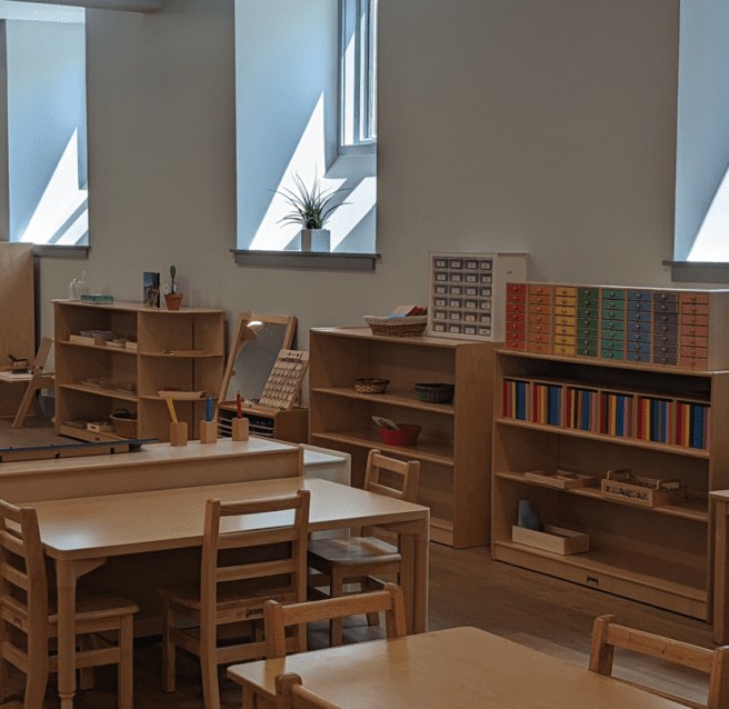 Tall windows over low wooden shelves of Montessori materials and child-sized tables