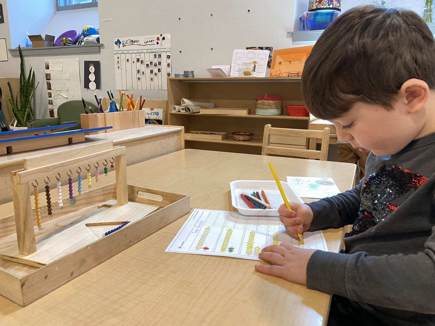 A boy concentrating on his writing work at a classroom table, with Montessori bead materials beside him
