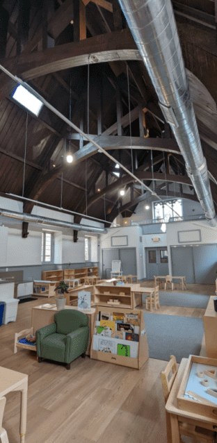 The main classroom under a high timber ceiling, with child-height shelves and a reading corner