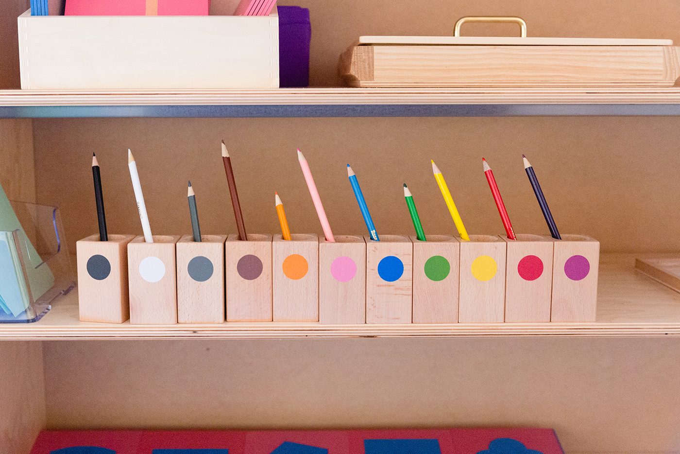 A row of wooden pencil holders, each a different colour, on a classroom shelf